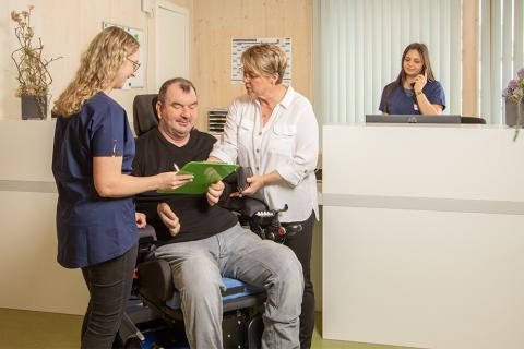 Zwei Mitarbeiterinnen des Medizinischen Zentrums für Erwachsene Menschen mit Behinderung (MZEB) sprechen mit einem Patienten, den im Rollstuhl sitzt. An der Rezeption telefoniert eine andere Dame.